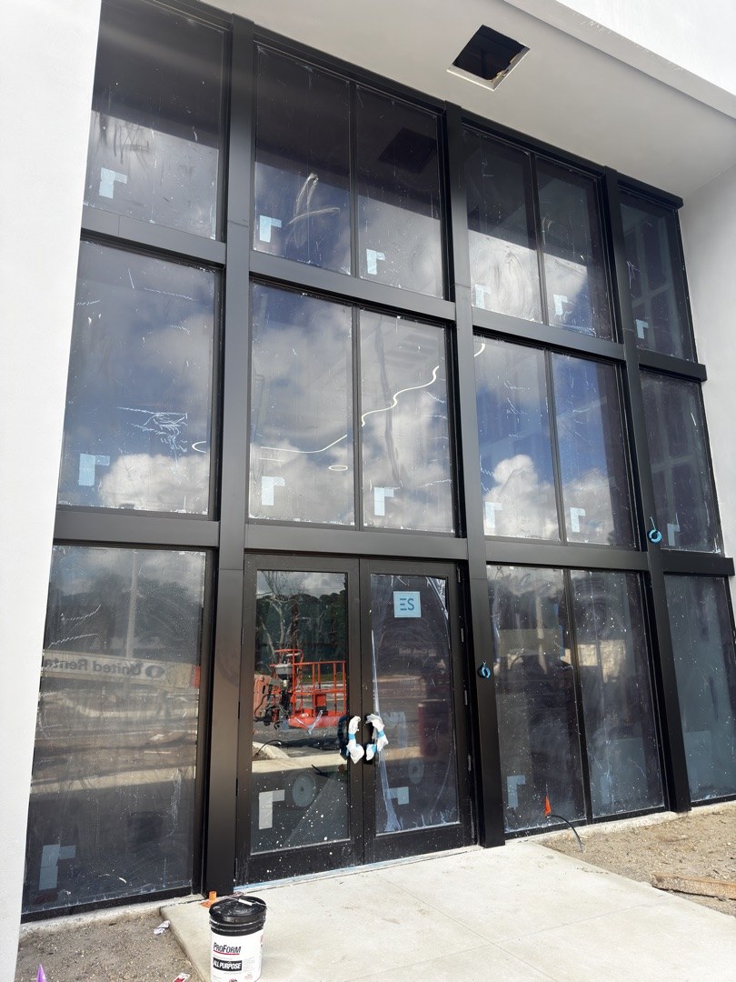KLUS Lighting two-story entrance storefront — ESWindows aluminum framing with impact glass installed, dark anodized finish, paired entry doors, protective film still on lites prior to handover