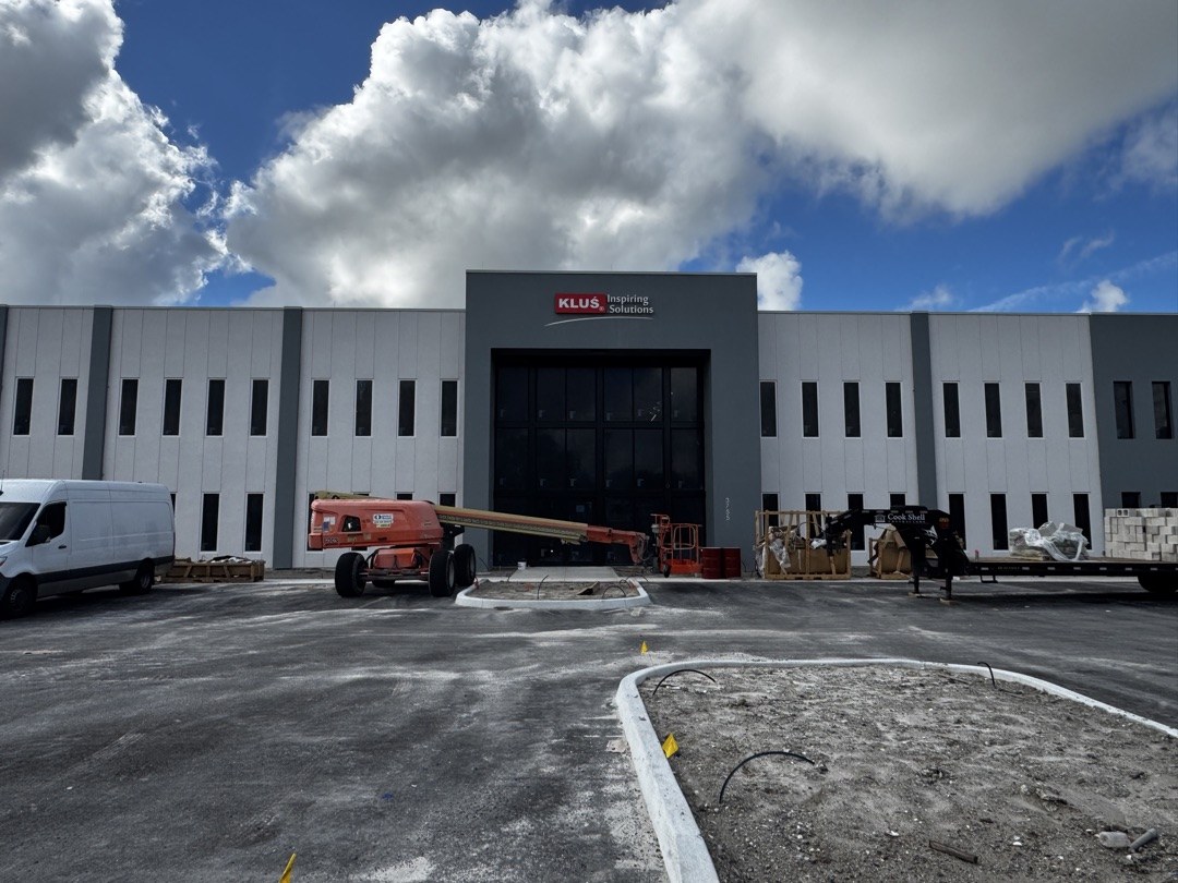 KLUS Lighting Vero Beach during construction — the full entry elevation with KLUS Inspiring Solutions signage installed above the two-story ESWindows storefront entrance, boom lift on site, Proctor Construction GC