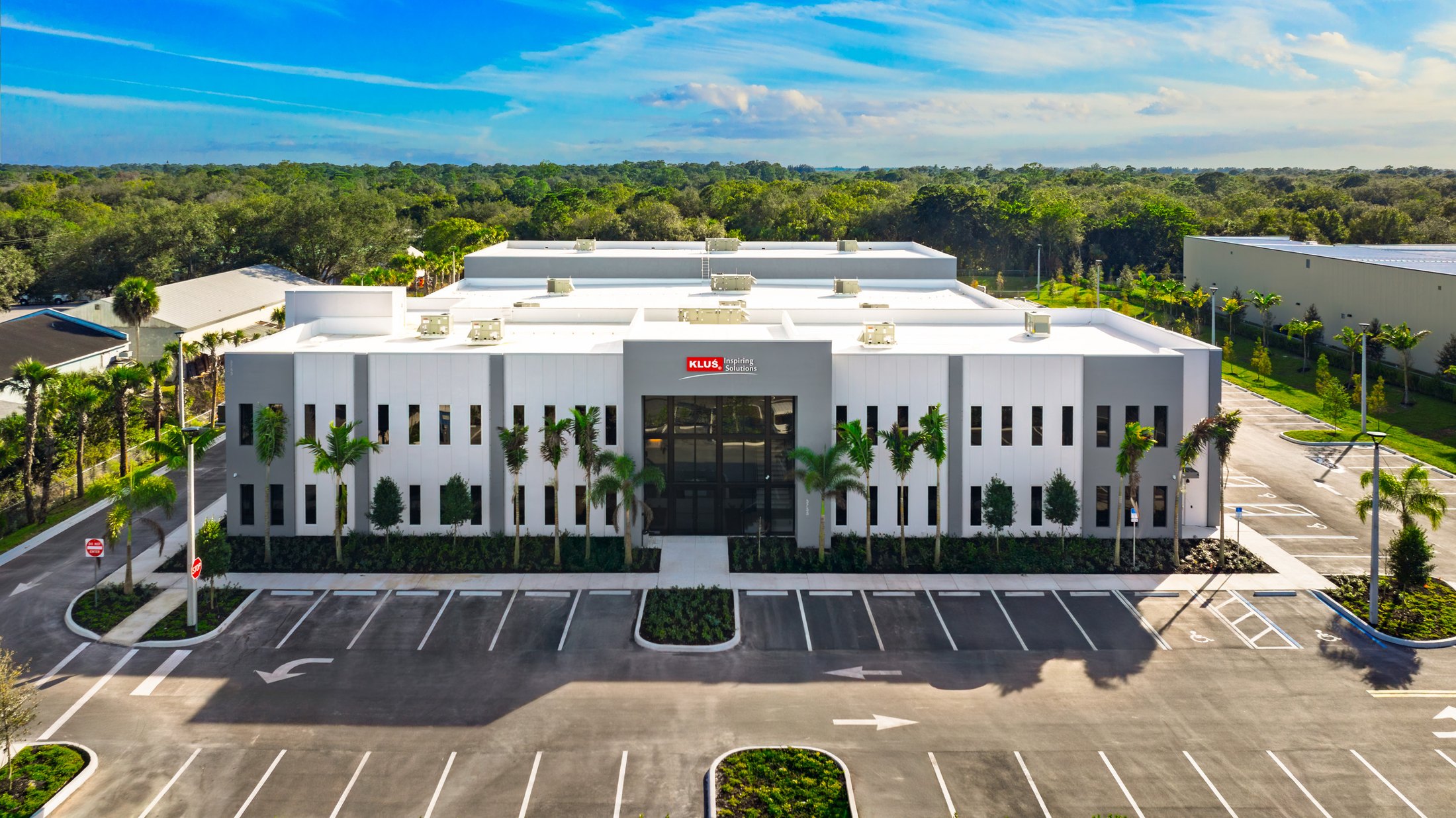 KLUS Lighting Vero Beach completed facility — wide aerial view of the front elevation with the KLUS Inspiring Solutions entry tower, palm landscaping, and the full ESWindows commercial storefront and window package by ACG, Proctor Construction GC
